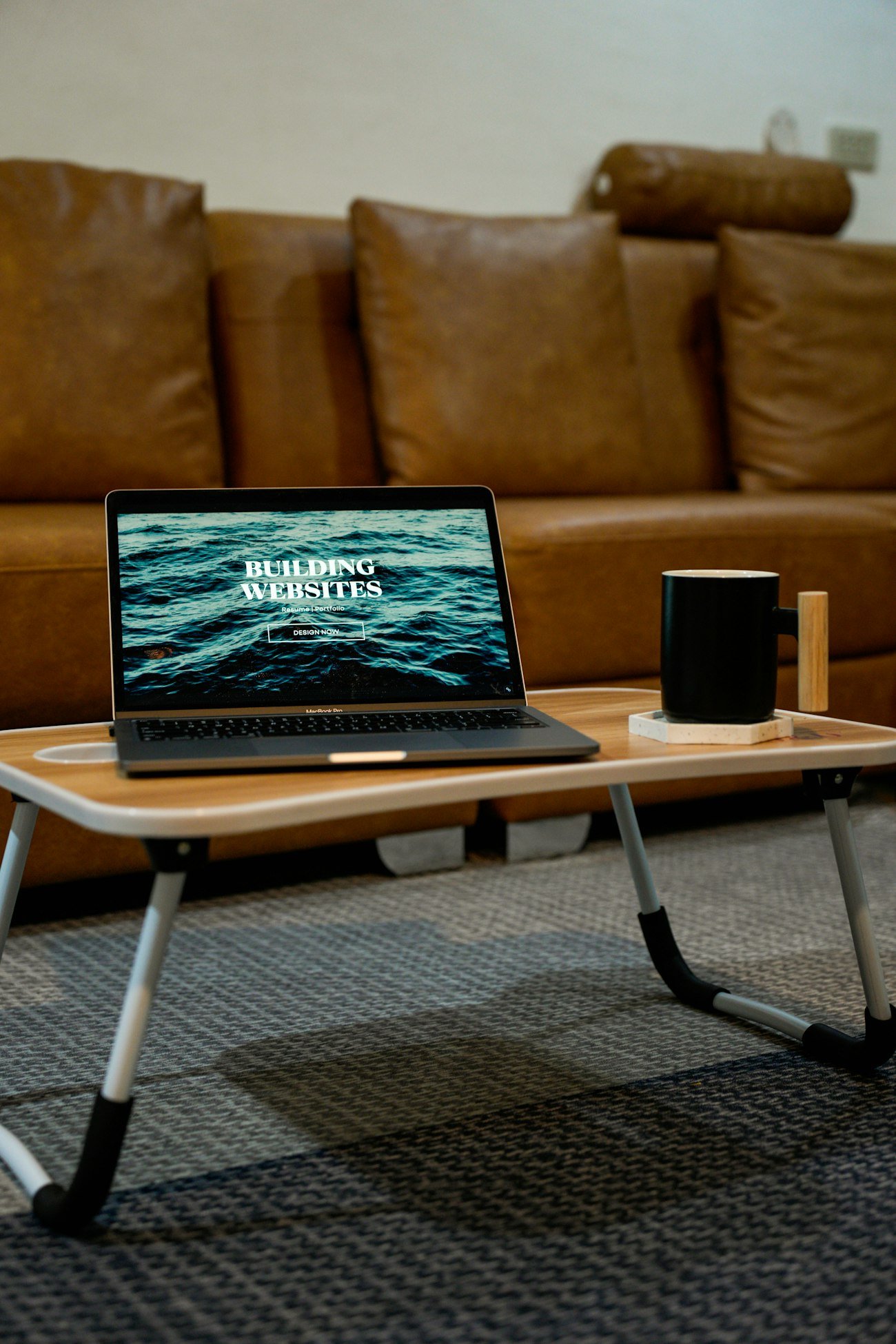 A laptop displaying the words 'Building Websites' rests on a small wooden table with black and white legs.